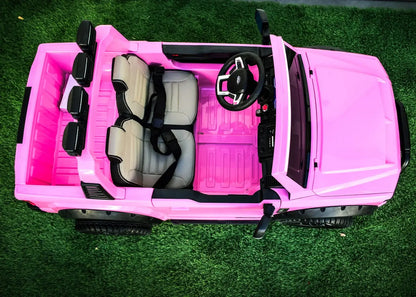 Kids ride-on Ford truck interior with dual seats, seat belts, and a vibrant pink exterior on grass.